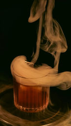 Smoky amber cocktail in a ribbed lowball glass with swirling smoke and a dark cherry garnish, dramatic dim bar-style lighting