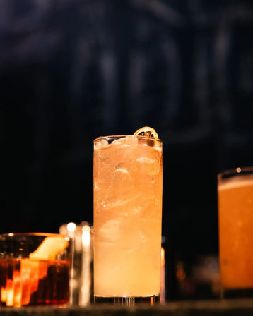 Sunset-hued highball cocktail with crushed ice and a dried citrus wheel garnish on the rim, glowing on a dark bar countertop with blurred drinks in the background.