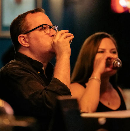 Close-up of two adults at a dimly lit bar taking shots — a man with glasses and a woman each raising a small glass in a lively nightlife scene.