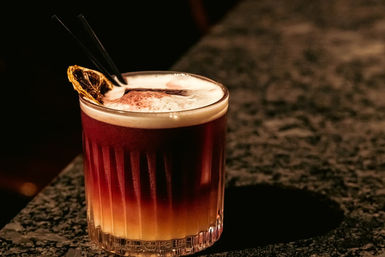 Two-tone layered cocktail with creamy foam, dried citrus wheel and two black straws in a short ribbed glass on a dark marble bar counter — moody bar nightlife vibe.