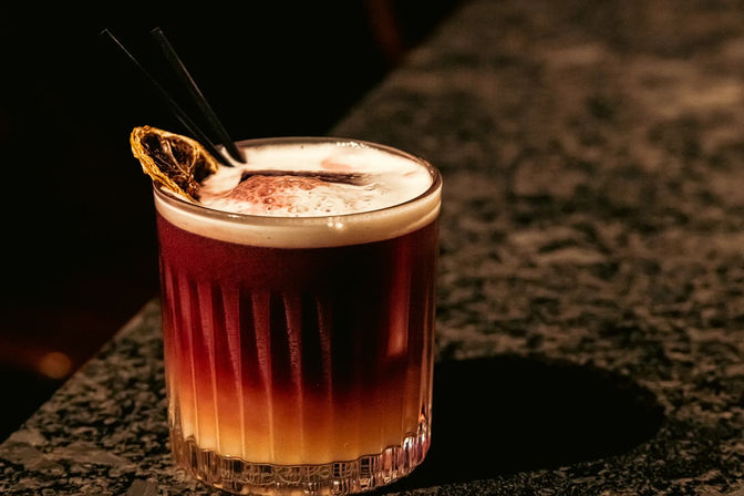 Two-tone layered cocktail with creamy foam, dried citrus wheel and two black straws in a short ribbed glass on a dark marble bar counter — moody bar nightlife vibe.