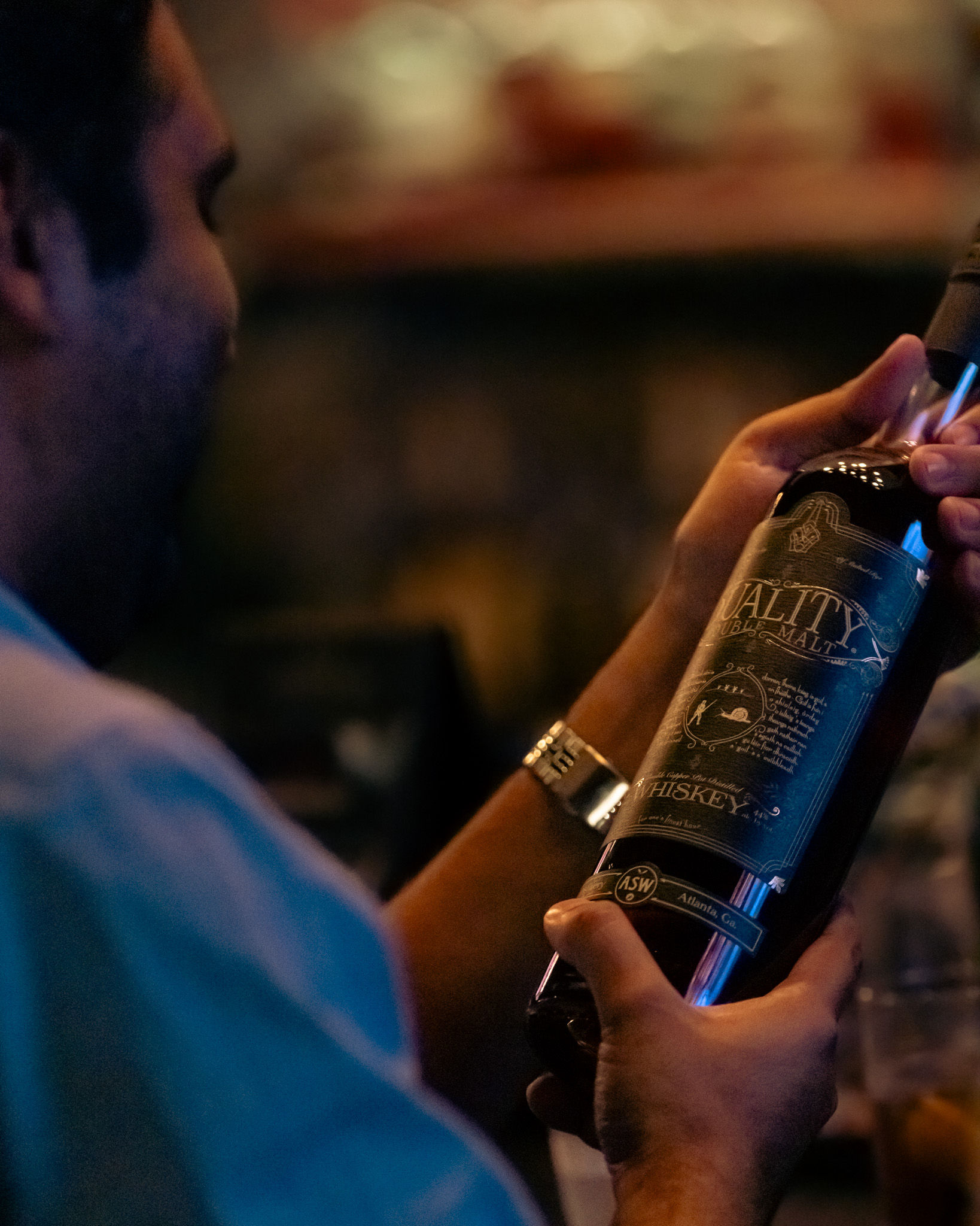 Close-up of hands holding a dark craft whiskey bottle and reading its ornate label in a dimly lit bar