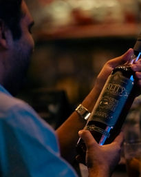 Close-up of hands holding a dark craft whiskey bottle and reading its ornate label in a dimly lit bar