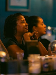 Profile of a person listening intently in a cozy, dimly lit bar or restaurant with hands under chin, blurred drinks and patrons in the warm evening interior — city nightlife vibe.