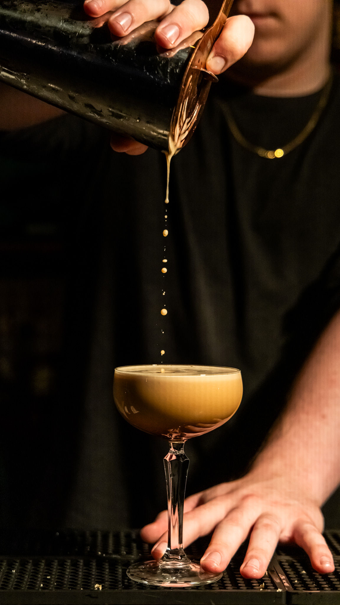 Bartender pouring a frothy espresso martini from a metal shaker into a coupe glass, golden droplets falling in a perfect vertical stream over a dimly lit bar.