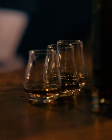 Three whiskey-tasting glasses with amber liquid lined up on a wooden bar counter, warm low-light and a blurred cozy background.