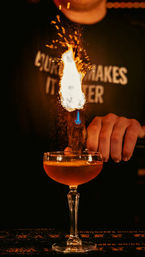 Close-up of a craft cocktail in a coupe glass as a bartender’s torch ignites orange zest, sending sparks and a tall flame above the drink in a dim bar.