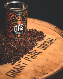 Tin of bourbon-infused coffee with ornate orange label on a wooden barrel top, surrounded by roasted coffee beans and warm moody lighting