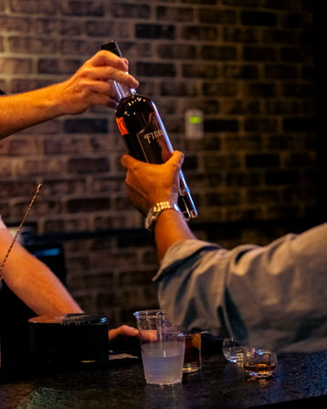 Two hands exchanging a whiskey bottle over a dim, brick-walled bar counter with plastic cups and shot glasses — lively nightlife scene.