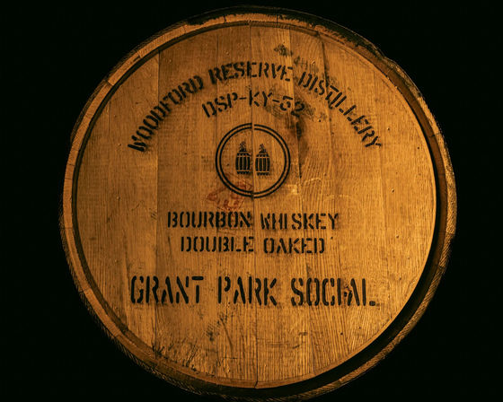Round oak bourbon barrel head stamped "Bourbon Whiskey Double Oaked" with distillery logo, warm amber lighting highlighting rustic wood grain against a black background.