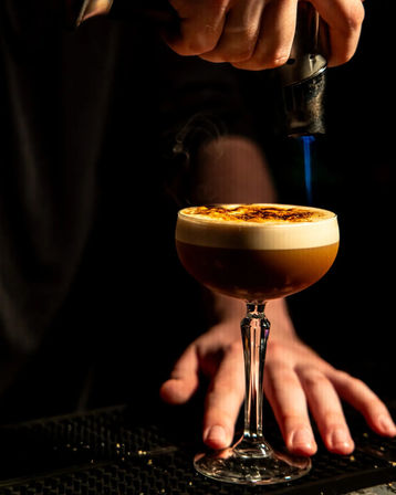 Close-up of a craft espresso cocktail in a coupe glass with creamy foam being caramelized by a blue torch flame at a dim bar