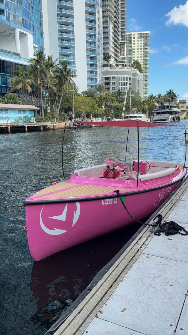 Bright pink leisure boat with canopy and floral centerpiece moored at a Florida waterfront canal, palm trees and high-rise condos in the background.