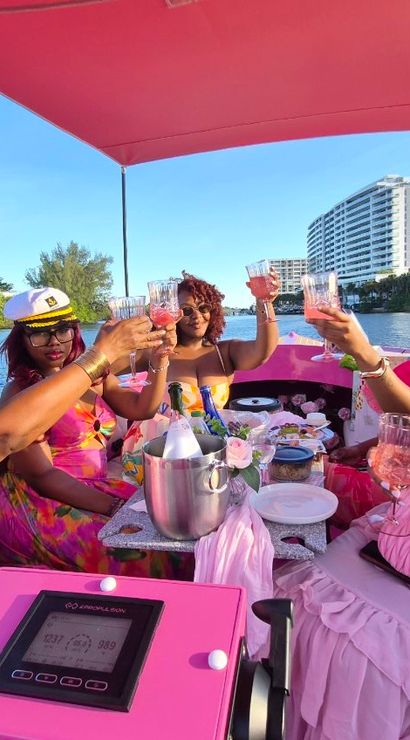 Group of five women in pink outfits toasting rosé on a pink party boat during a sunny waterfront brunch with high-rise condos along the shore