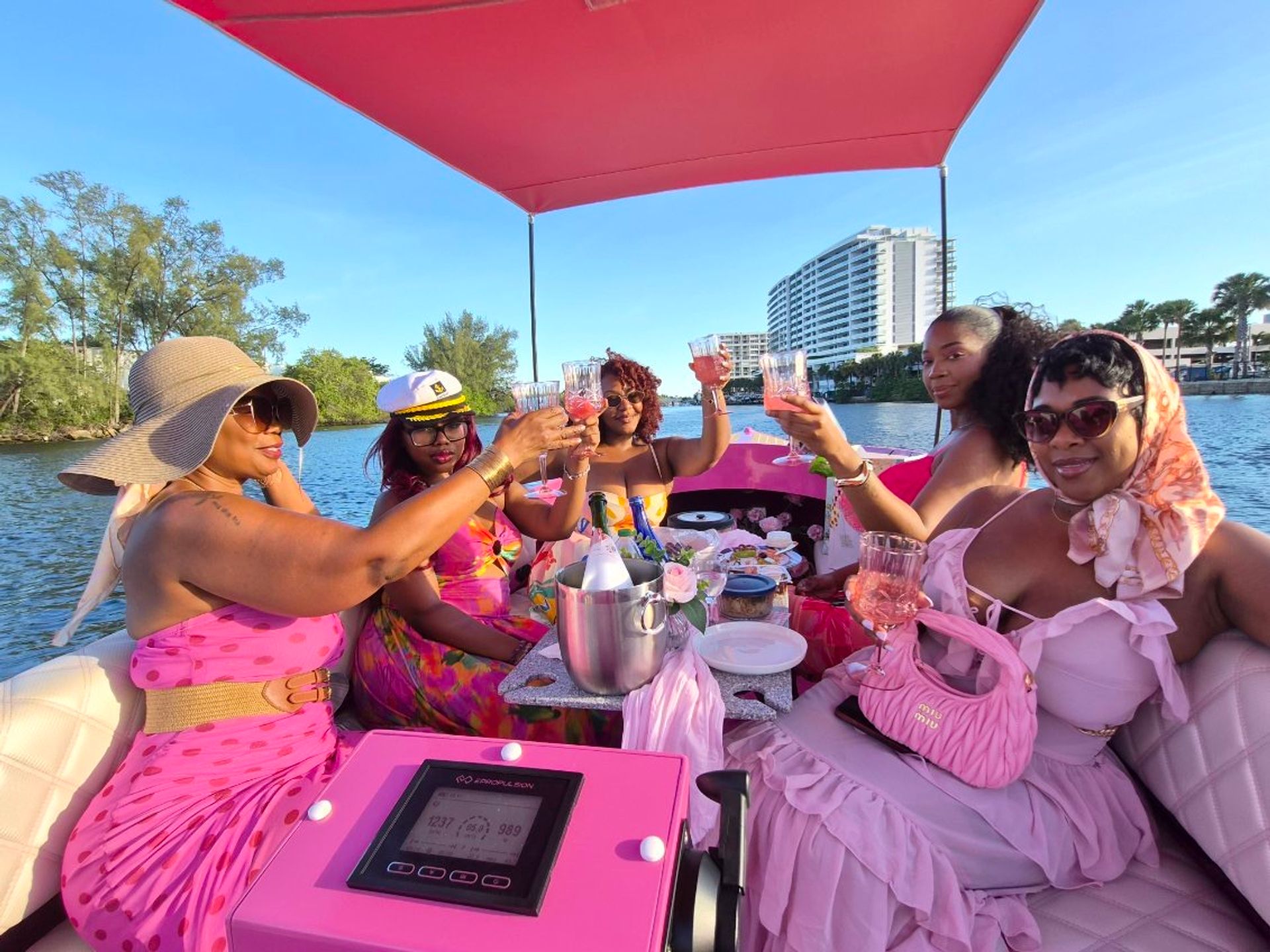 Group of five women in pink outfits toasting rosé on a pink party boat during a sunny waterfront brunch with high-rise condos along the shore