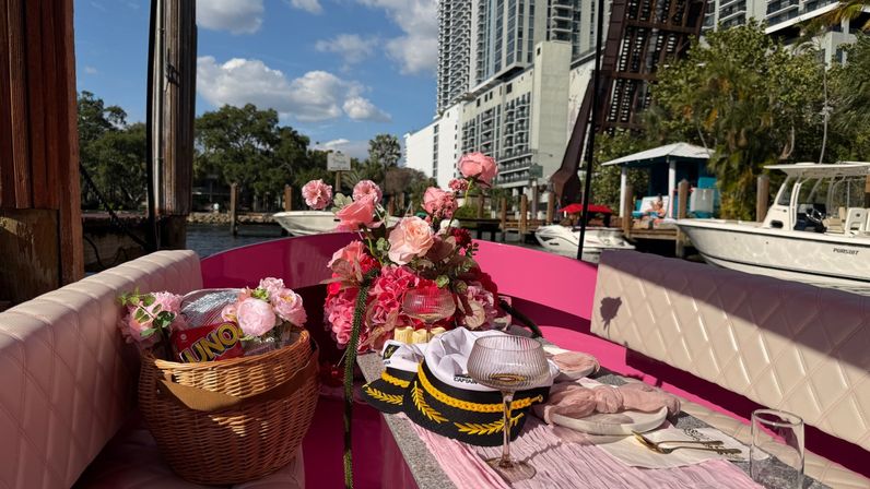 Cheerful pink boat interior staged for a waterfront picnic with quilted seats, wicker basket (UNO cards), pink floral centerpiece, captain’s hats and glassware, docked at an urban marina with boats and high-rise buildings in the background.