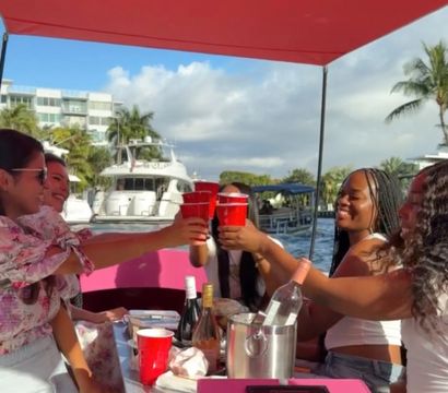 Friends toasting with red cups on a bright pink party boat under a red canopy at a sunny marina, yachts and palm trees in the background.