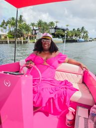 Smiling woman in a bright pink dress and captain's hat steering a vibrant pink boat on a palm-lined waterfront