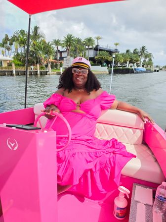 Smiling woman in a bright pink dress and captain's hat steering a vibrant pink boat on a palm-lined waterfront