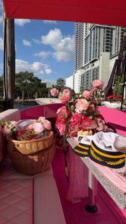 Bright pink boat at a marina with a pink floral centerpiece, wicker picnic basket holding a card game, captain hats and wine glasses on a small table, with waterfront docks and city high-rise skyline under a blue sky.