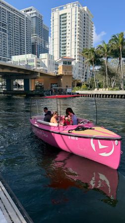 Bright pink leisure boat with passengers and a floral bouquet cruising a sunny Florida urban canal beneath a bridge, with palm trees and high-rise waterfront condos in the background.