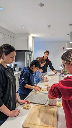 Group baking session in a bright modern shared kitchen: five people shaping braided bread rolls on a white island countertop with baking trays, stainless refrigerator, and pendant lights.