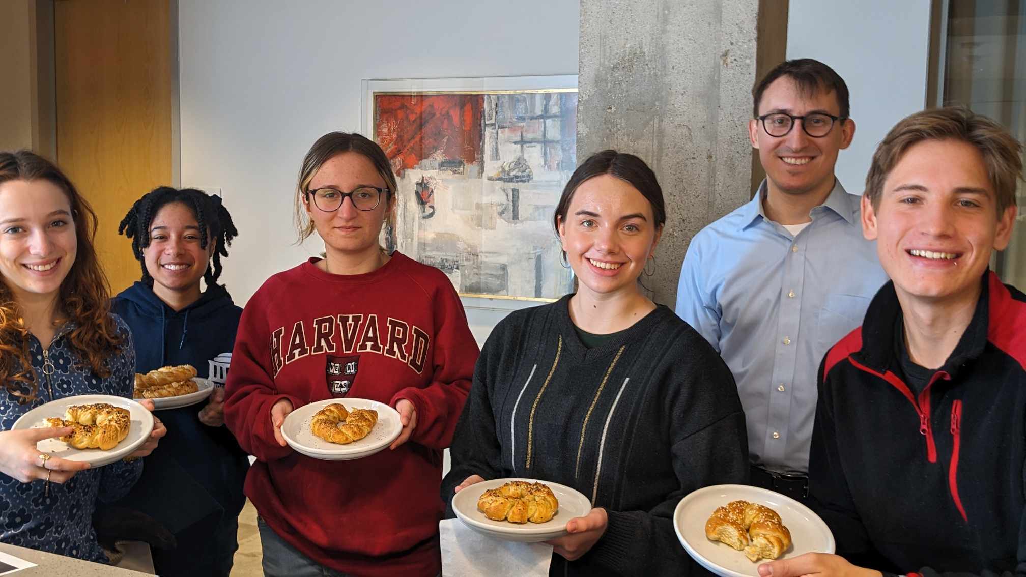 Office team breakfast: six smiling coworkers in a modern office holding plates with bagels and pastries, casual clothing, concrete column and abstract painting in the background.