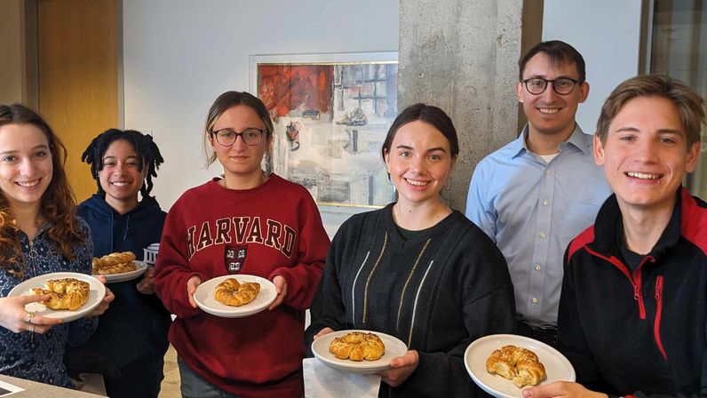 Office team breakfast: six smiling coworkers in a modern office holding plates with bagels and pastries, casual clothing, concrete column and abstract painting in the background.