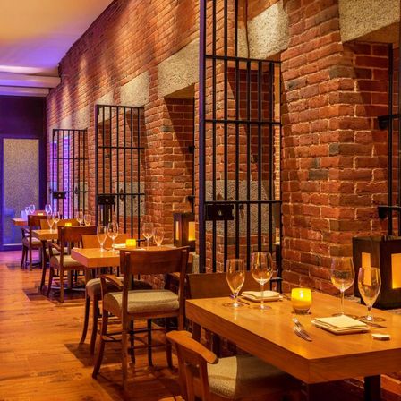 Cozy candlelit restaurant interior in a historic red-brick space, intimate wooden tables set with wine glasses and napkins along an exposed brick wall framed by black metal grilles