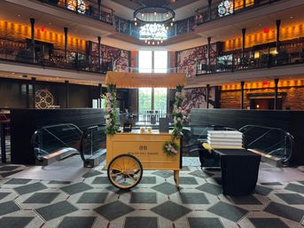 Yellow flower‑adorned bakery cart centered in a grand multi-level hotel atrium with glass escalators, balcony seating, geometric carpet pattern, and a large chandelier overhead.