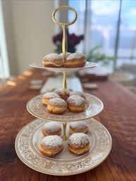 Three-tier gold-handled serving stand holding powdered-sugar doughnuts on ornate vintage china plates, arranged on a wooden dining table in soft natural light