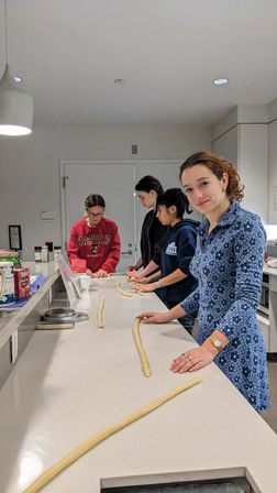Group of four people in a bright modern communal kitchen rolling long ropes of dough along a white island countertop during a casual baking session, one person in a blue floral dress smiling at the camera.