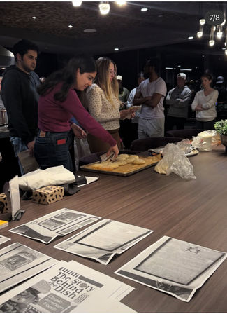 Group of people gathered around a modern kitchen island during a casual cooking class, a woman serving freshly baked bagels from a tray while printed recipe handouts and ingredients sit on the counter.