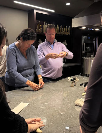 Adults shaping dough at a modern home kitchen island, marble countertop with a large pot on the stove and spice jars nearby during a casual group cooking activity