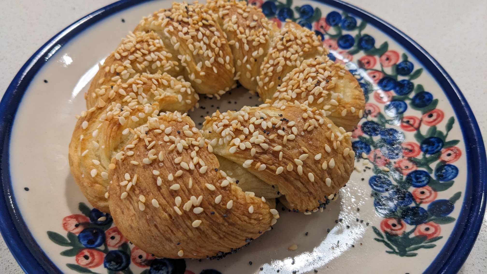 Golden sesame-topped braided pastry ring on a hand-painted floral ceramic plate, close-up on a white countertop