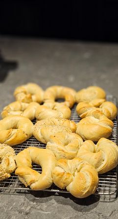 Golden fresh-baked bagels and twisted rolls, some sesame-topped, cooling on a wire rack on a kitchen countertop.