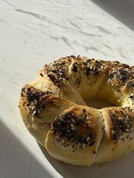 Sunlit golden twisted everything bagel sprinkled with sesame, poppy seeds and onion flakes on a white textured cloth — artisanal breakfast pastry.