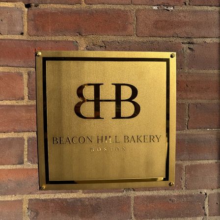 Gold metal plaque with a stylized double‑B monogram and 'bakery' lettering mounted on a red brick wall outside a Boston storefront.