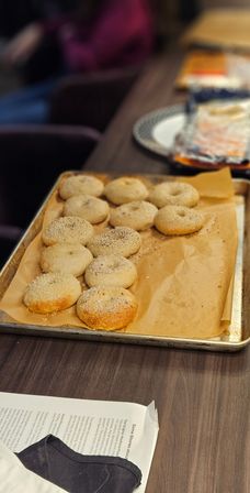 Tray of freshly baked plain and seeded bagels with golden crusts on a parchment-lined baking sheet resting on a dark wood table, plate and papers blurred in the background.
