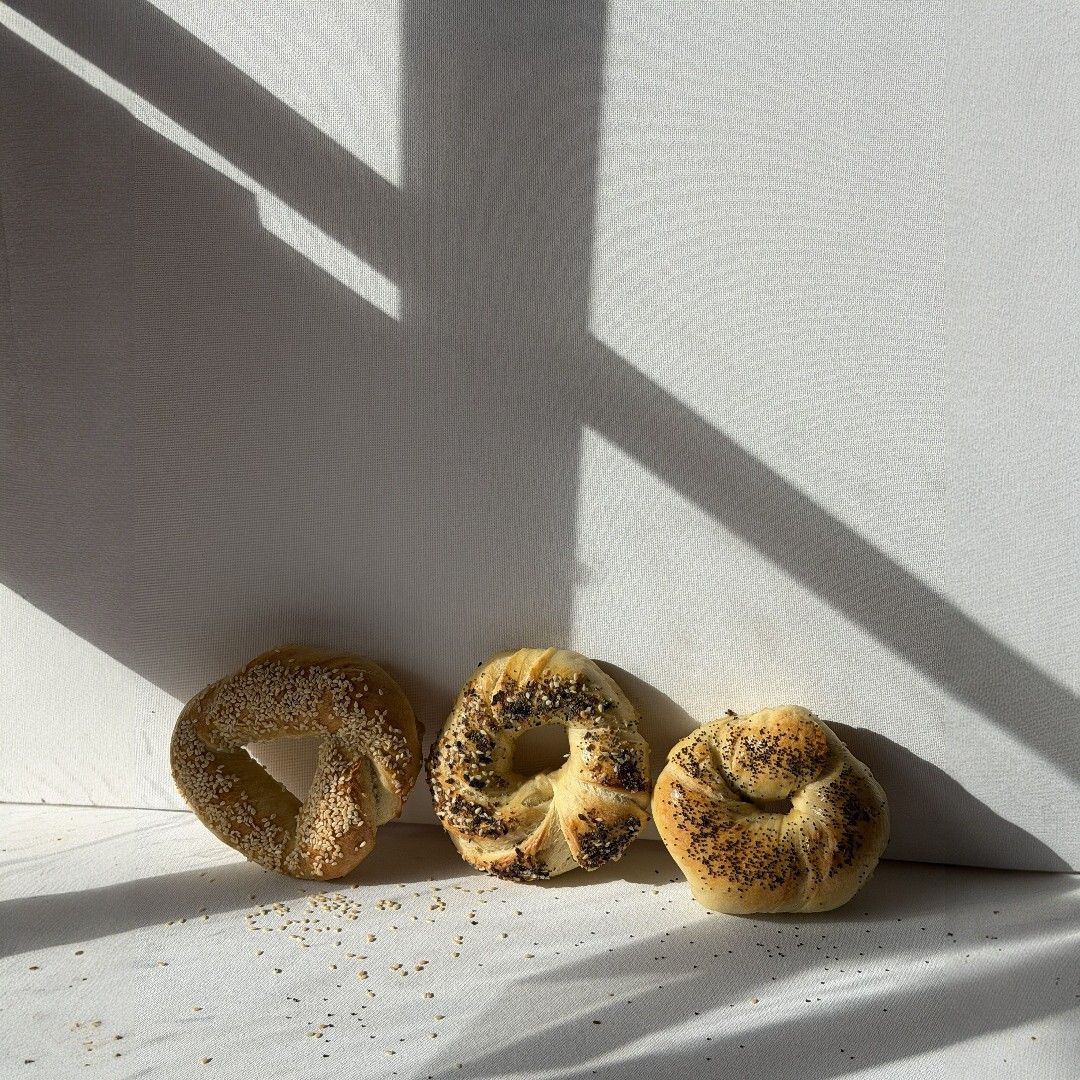 Three seeded bagels (sesame and poppy) basking in slanted window light on a white surface, minimalist breakfast scene with scattered seeds
