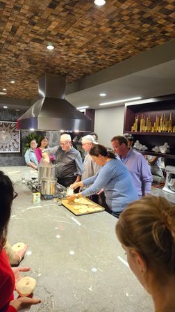 Group cooking class at a modern kitchen island: people shaping bagels beside a large pot under a stainless steel range hood, marble countertop and wood-tile ceiling.
