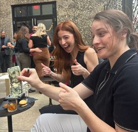 Two women laughing and giving thumbs up on an outdoor patio bar; one holds a crumpled dollar bill above a small table with a cocktail and mini burger, lively crowd in the background.