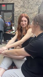 Two women chatting at an outdoor patio event — a red‑haired woman in a black sleeveless top smiles across to her companion, seated casually with a textured stone wall, doorway, and event tables in the background.