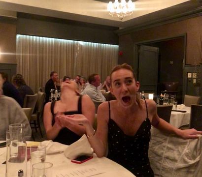 Two young women animatedly reacting at a round table in a dimly lit restaurant dining room, with menus, glasses and a lemon on the table and other diners and a chandelier in the background.