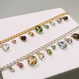 Close-up jewelry display of gold and silver charm bracelets with sparkling bezel-set multicolored gemstone teardrop and round charms in green, blue, purple, red, yellow and clear tones.