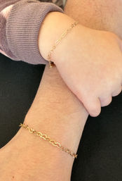 Close-up of an adult and child wrist wearing matching delicate gold heart-link chain bracelets, child in a purple cuff touching the adult wrist against a black background.