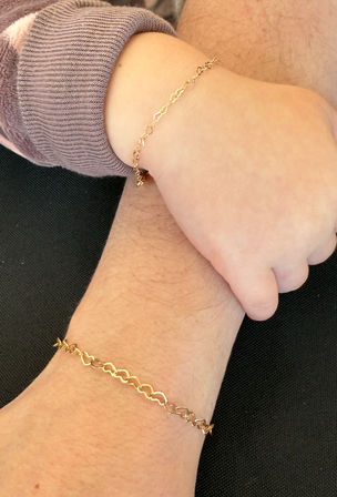 Close-up of an adult and child wrist wearing matching delicate gold heart-link chain bracelets, child in a purple cuff touching the adult wrist against a black background.