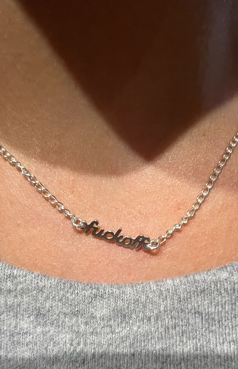 Close-up of a silver script nameplate necklace resting on a collarbone, shiny chain and cursive word pendant above a gray knit top