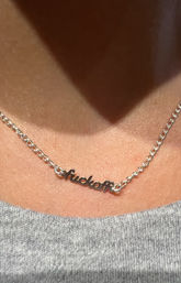 Close-up of a silver script nameplate necklace resting on a collarbone, shiny chain and cursive word pendant above a gray knit top