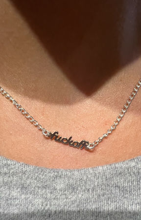 Close-up of a silver script nameplate necklace resting on a collarbone, shiny chain and cursive word pendant above a gray knit top