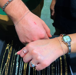 Close-up of two hands touching over a jewelry display, featuring silver chain bracelets, a vintage-style wristwatch, heart and gemstone rings, and rows of sparkling gold and silver chains beneath.